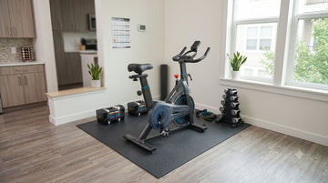 Un aménagement complet et abordable de salle de sport pour débutants, avec un vélo Wenoker JC302, des poids réglables et un tapis en PVC dense dans le coin d'un petit appartement